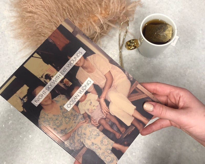 Hand holding Night Sky with Exit Wounds by Ocean Vuong with tea and fuzzy pillow