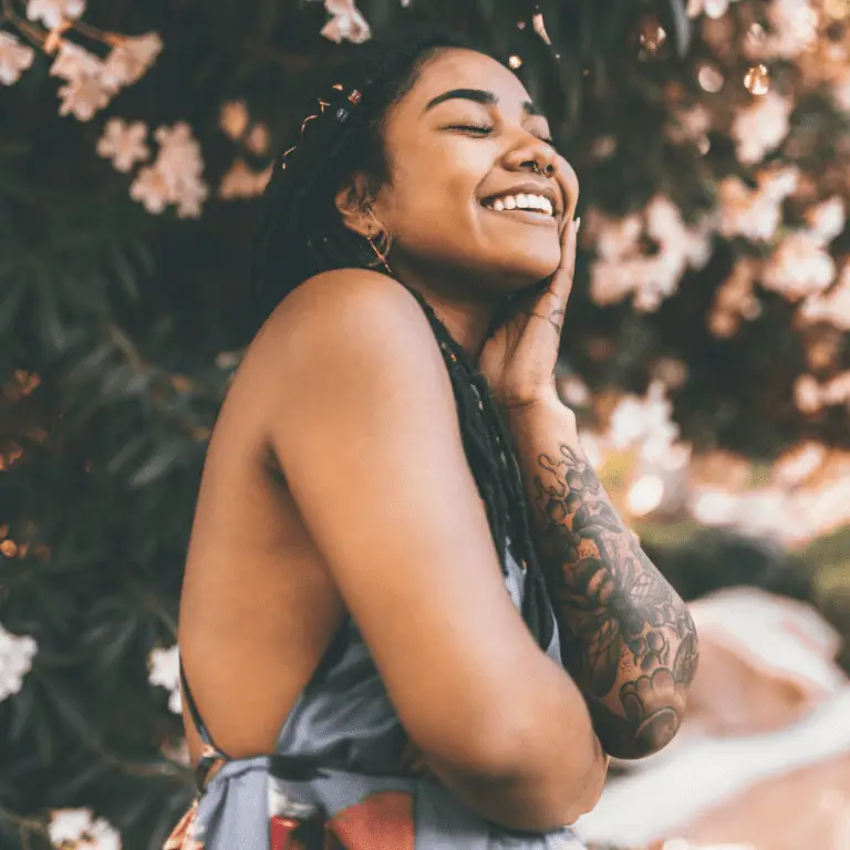 Black woman standing in front of a flowering tree with her arm wrapped around her waist smiling with pure joy