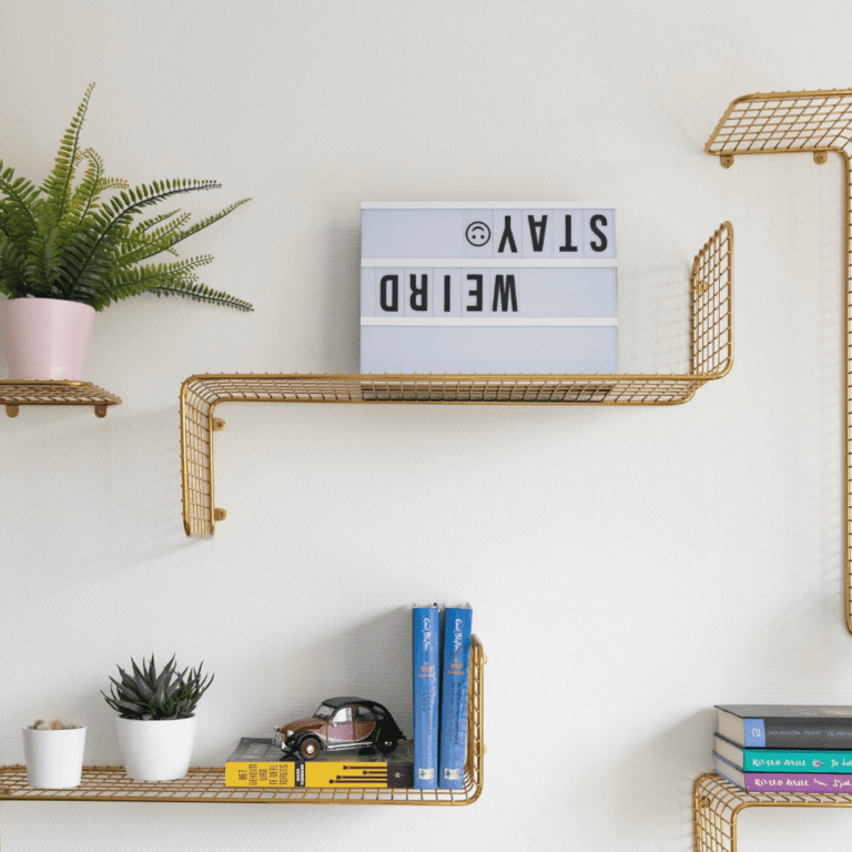 Gold wire shelves on a white wall. One shelf has an upside-down sign that says "stay weird".