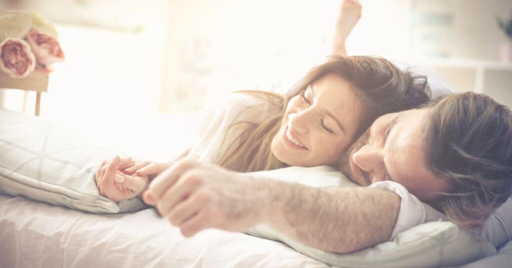 Couple smiling and holding hands, lying together on a bed in a bright, cozy room.