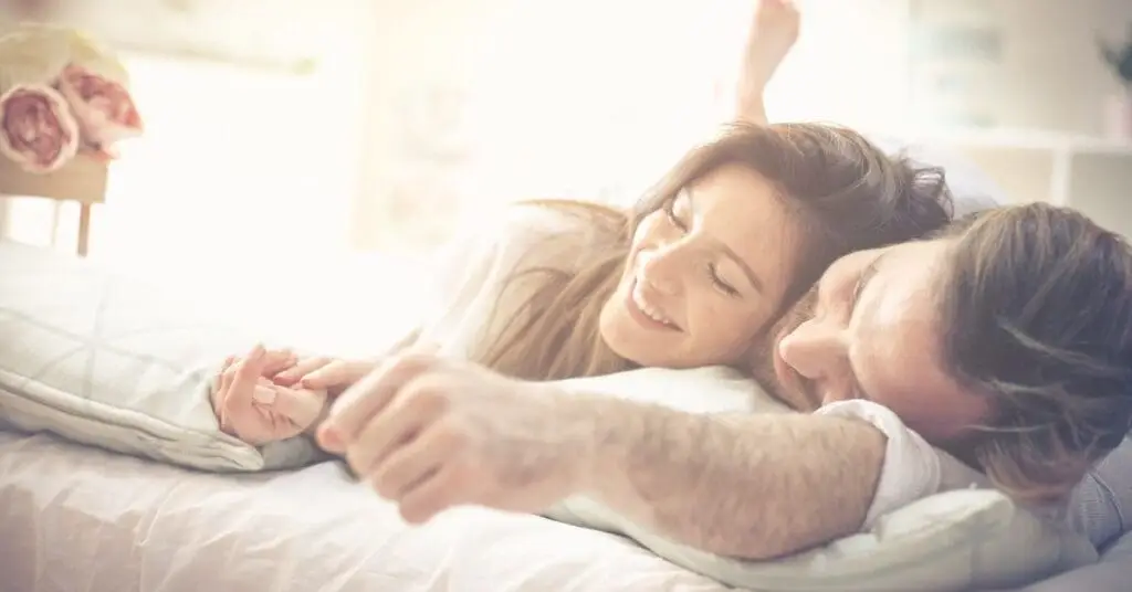 Couple smiling and holding hands, lying together on a bed in a bright, cozy room.