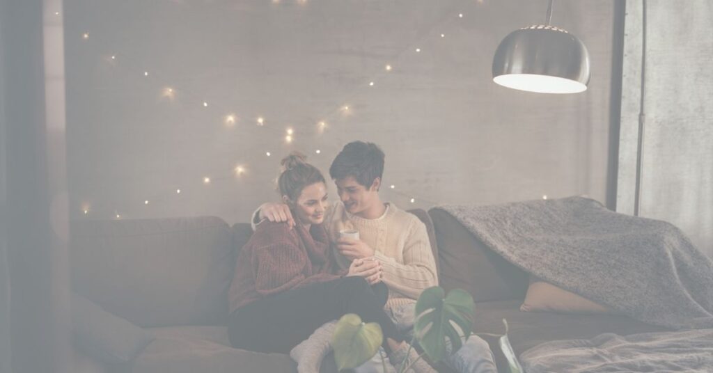 A couple sits close together on a cozy couch, sharing a warm drink under string lights.