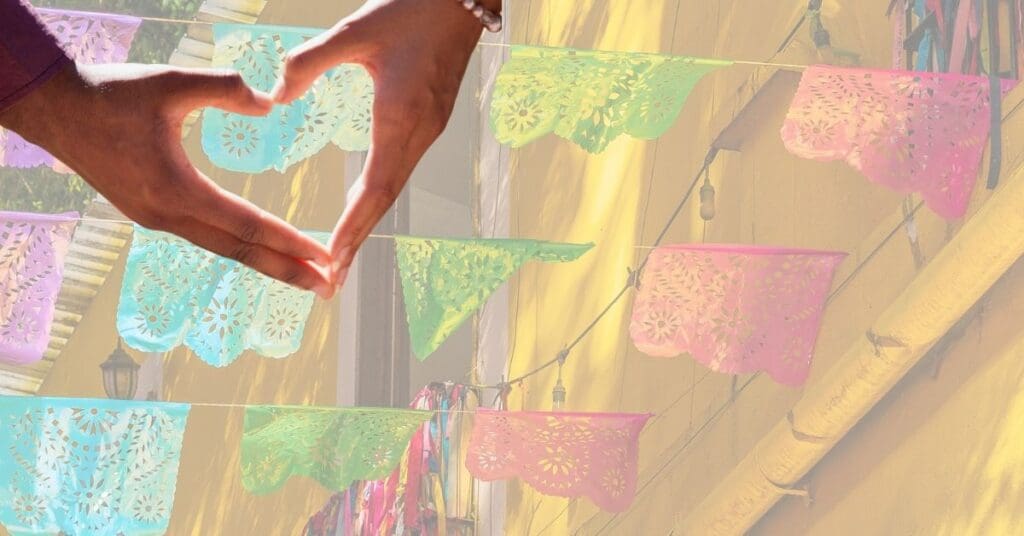 Hands form a heart in front of colorful papel picado hanging on a yellow building.