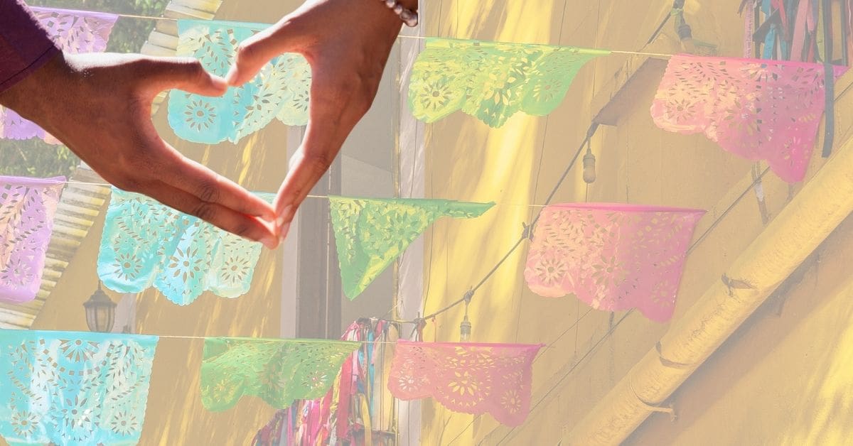 Hands form a heart in front of colorful papel picado hanging on a yellow building.