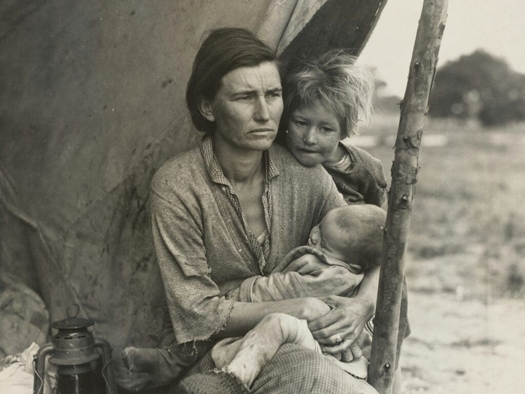 Sepia old photo of a woman holding her infant child with another young child standing behind her outside a cloth tent held up with a long stick.