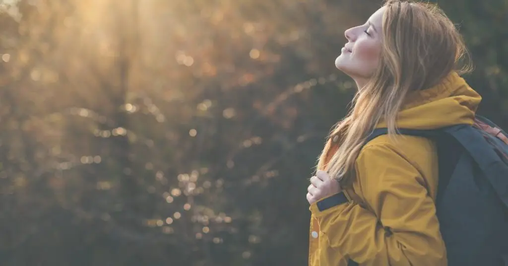 woman in jacket with backback enjoying nature and lifting her face to the sun while hiking through the woods