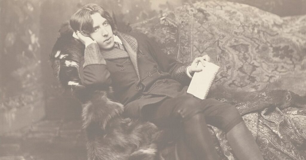 Author Oscar Wilde, well dressed, reclining on a fur-covered chair with a journal in his hand