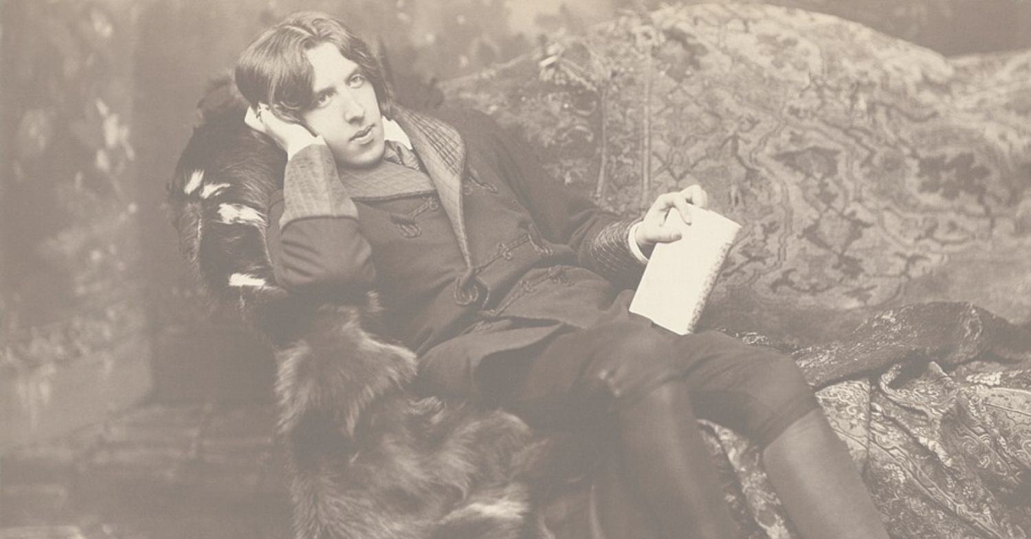Author Oscar Wilde, well dressed, reclining on a fur-covered chair with a journal in his hand